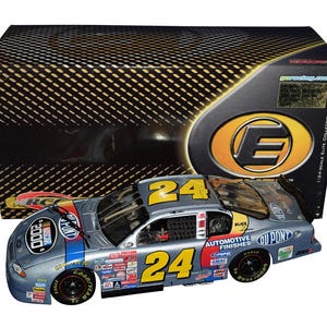 Op de afbeelding: Een zilver en blauw NASCAR-raceauto model met het nummer 24 in geel. De auto heeft verschillende sponsorstickers en wordt gepresenteerd voor de originele doos. De doos heeft een zwart-gouden patroon en het logo van de fabrikant.