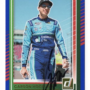 May include: A rectangular sports trading card featuring a photograph of a race car driver wearing a light blue and dark blue racing suit, a black cap, and sunglasses. The card has a blue border, green accents, the text Carson Hocevar, and a black ink autograph.
