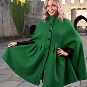 Op de afbeelding: Een felgroene cape met een hoge kraag en knoopsluiting. De cape heeft een geplooid ontwerp en valt tot halverwege de dij. De persoon draagt er een zwarte top onder. De achtergrond toont een stenen kasteel.