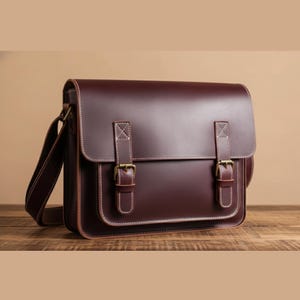 May include: A burgundy leather messenger bag. The bag has a rectangular shape, a flap closure with two buckle straps, white stitching, and a shoulder strap. The bag is on a wooden surface.