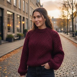 Puede incluir: Un suéter de color burdeos con un patrón de punto texturizado. El suéter tiene cuello redondo y mangas largas. La persona de la imagen lleva el suéter en una calle adoquinada.