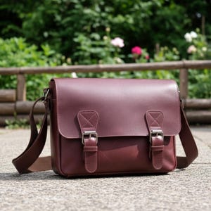 May include: A burgundy leather messenger bag with a flap closure and two silver buckles. The bag has a shoulder strap and is sitting on a concrete surface. The background features greenery and a wooden fence.