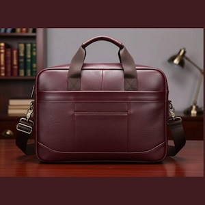 May include: A burgundy leather briefcase with a top handle and a detachable shoulder strap. The bag has a front pocket and metal hardware. The briefcase is sitting on a wooden surface.