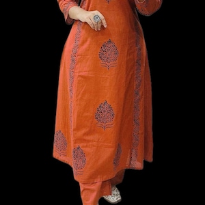 Può includere: Completo kurta e pantaloni abbinati color ruggine, con maniche a tre quarti. Il kurta presenta un motivo ad albero blu scuro e dettagli simili sui lati. L'outfit è indossato con ballerine decorate.