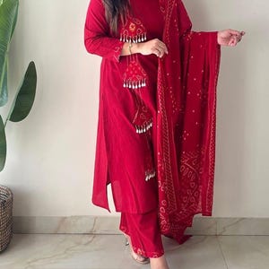 Puede incluir: Un vibrante conjunto de salwar kameez rojo. El conjunto incluye una túnica de manga larga con borlas decorativas, pantalones a juego y una bufanda estampada. El atuendo está hecho de una tela ligera, adecuada para climas cálidos.