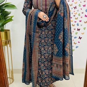 Może przedstawiać: Ciemnoniebieski indyjski zestaw salwar kameez z pasującą dupattą. Str&oacute;j ma skomplikowane wzory kwiatowe i geometryczne w odcieniach czerwieni, brązu i bieli. Top z długim rękawem ma dekolt w kształcie dziurki od klucza.