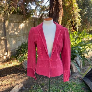 May include: A red suede blazer with decorative stitching along the seams and cuffs. The blazer is displayed on a mannequin, showcasing its tailored fit and classic design. The jacket has a single button closure.