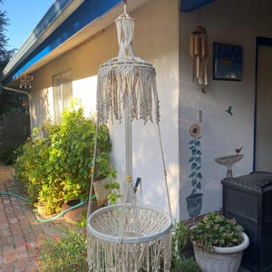 May include: A cream-colored macrame hanging chair with a circular seat and a fringed canopy. The chair is suspended from a beam on a porch, surrounded by greenery and a brick walkway. The chair is made of woven cotton.