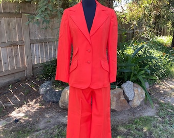 May include: A vibrant red suit, consisting of a blazer and wide-leg pants, displayed on a mannequin. The blazer features a two-button closure and two front pockets. The suit is made of a textured fabric and is set against a natural outdoor backdrop.