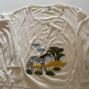 May include: Cream-colored long-sleeved shirt with a crew neck. The shirt features an embroidered design of a zebra, trees, and clouds. The zebra has black and gray stripes, and the trees are green.