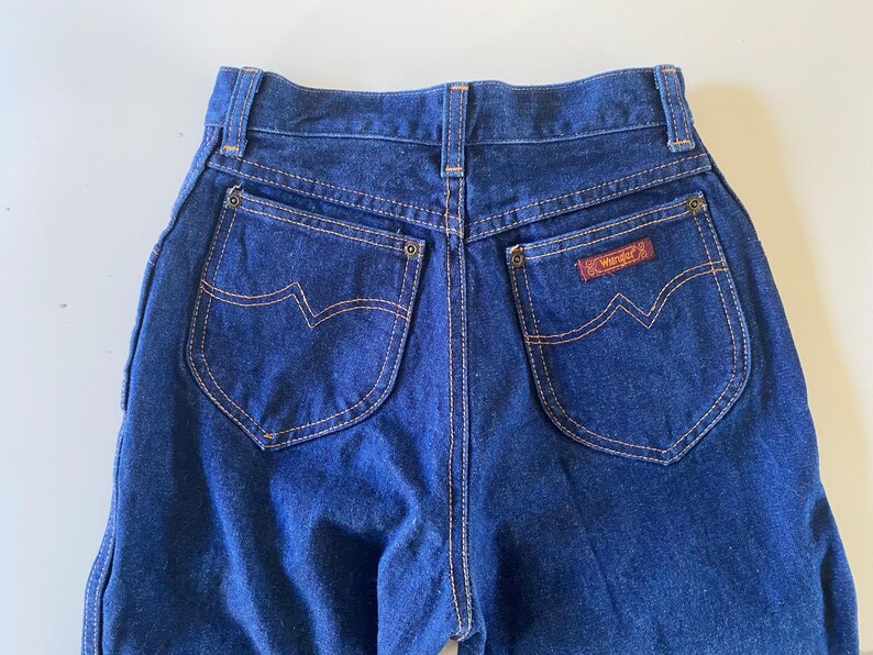 May include: A pair of dark blue denim jeans with a brown leather patch that says "Wrangler" on the back right pocket.