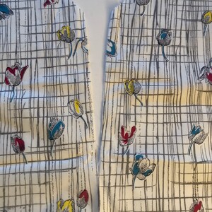 May include: Two pieces of white fabric with a grid pattern and a floral design of blue, yellow, and red tulips. The fabric is cut along the edge, ready to be sewn together.
