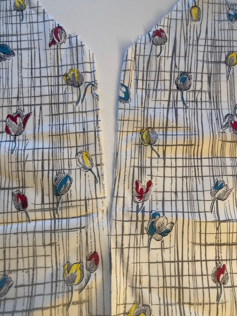 May include: Two pieces of white fabric with a grid pattern and a floral design of blue, yellow, and red tulips. The fabric is cut along the edge, ready to be sewn together.