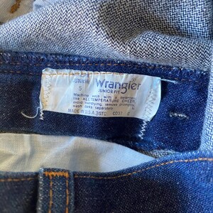 May include: A blue denim Wrangler Junior Fit jean label with white stitching. The label has the text "JUNIOR" and "5" printed on it. The label also has washing instructions: "Machine wash with a detergent like ALL TEMPERATURE CHEER, avoid overdrying, remove promptly, wash darks separately." The label also has the text "MADE IN U.S.A. 3GTC C037 C".