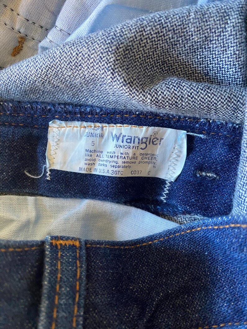 May include: A blue denim Wrangler Junior Fit jean label with white stitching. The label has the text "JUNIOR" and "5" printed on it. The label also has washing instructions: "Machine wash with a detergent like ALL TEMPERATURE CHEER, avoid overdrying, remove promptly, wash darks separately." The label also has the text "MADE IN U.S.A. 3GTC C037 C".