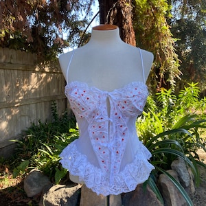 May include: White lingerie top with red heart pattern and lace trim. The top features spaghetti straps and a ruffled neckline and hem. The garment is displayed on a mannequin in an outdoor setting with greenery in the background.