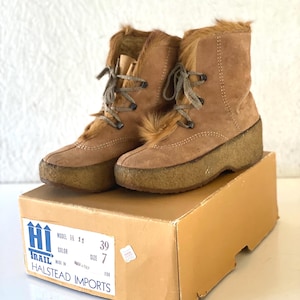 May include: A pair of brown suede boots with faux fur trim and laces. The boots are size 7 and sit on a platform sole. The boots are displayed on a cardboard box with the text "Halstead Imports".