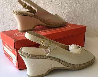 May include: A pair of beige canvas wedge sandals with a slingback strap and a buckle closure. The sandals are sitting on a red box with the word "Brown Shoe" printed on it.