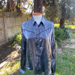 May include: A long-sleeved, button-down shirt in a shimmering, dark gray fabric. The shirt features a classic collar, buttoned cuffs, and black trim along the yoke and seams. The shirt is displayed on a mannequin outdoors.