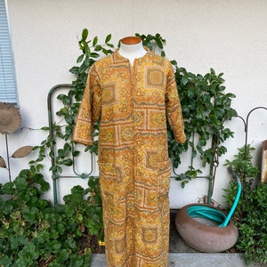 May include: A long, quilted robe with a geometric pattern in shades of brown, yellow, and green. The robe has a zipper closure and long sleeves.