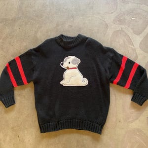 May include: Black knitted sweater with red stripes on the sleeves and a white embroidered dog applique on the front.