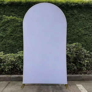 May include: A white arch backdrop with a rounded top, supported by gold-colored metal legs. The backdrop is made of a smooth, solid white fabric. The background features green foliage.