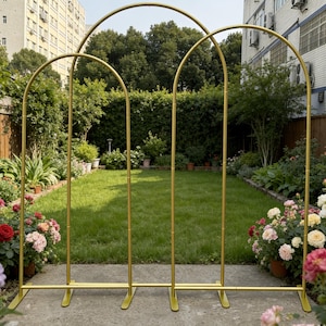 Op de afbeelding: Een gouden metalen trouwboog met drie gebogen secties. De boog is buiten opgesteld in een tuin met groen gras, bloemen en bomen. De boog is ontworpen voor bruiloften en andere buitenevenementen.