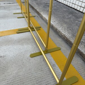 May include: A gold metal backdrop stand with a rectangular base and vertical poles. The stand is designed for events and displays. The stand is on a concrete surface with yellow painted lines.