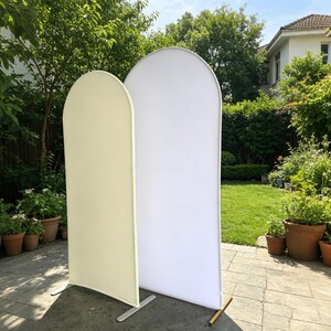 May include: Two arched backdrop stands, one cream and one white, are displayed outdoors. The fabric panels are stretched over metal frames, with a curved top and rectangular base. The stands are positioned on a patio with a green lawn and trees in the background.