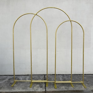 May include: A gold metal arch backdrop with three connected sections. Each section has a curved top and vertical supports, creating an arch shape. The backdrop is designed for events and celebrations.