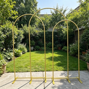 May include: Three gold metal arch wedding backdrops stand on a concrete patio in a backyard. The arches are arranged in a row, with a curved top and rectangular base. Lush green grass and foliage fill the background.