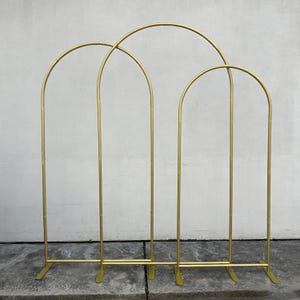 May include: A gold metal arch backdrop with three connected sections. Each section has a curved top and vertical supports, creating an arch shape. The backdrop is designed for events and celebrations.