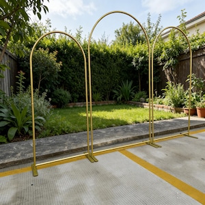 May include: Three gold metal arch wedding decorations stand outdoors on a concrete surface. Each arch has a rounded top and is connected by a horizontal bar at the base. The arches are in front of a green lawn and trees.