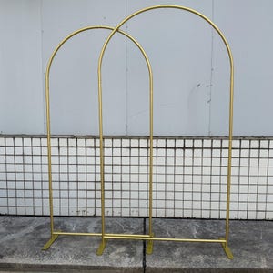 May include: Two gold metal arch wedding backdrops. Each arch has a rounded top and is supported by vertical poles and a rectangular base. The arches are designed for decorative purposes, such as weddings or events.