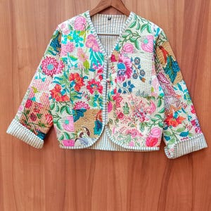 Könnte beinhalten: Gesteppte Jacke mit floralem Patchwork-Design. Die Jacke hat einen V-Ausschnitt, drei Knöpfe und gestreifte Manschetten. Das Blumenmuster umfasst rosa, orange und blaue Blumen auf cremefarbenem Hintergrund. Auf einem Holzbügel aufgehängt.