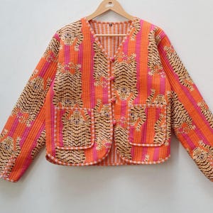 Puede incluir: Chaqueta acolchada con estampado de rayas de tigre en naranja, rosa y beige. La chaqueta tiene cuello en V, dos bolsillos delanteros y cierre de botones. La prenda se exhibe en una percha de madera.