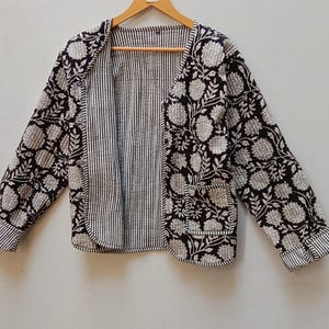 May include: A black and white quilted jacket with a floral pattern. The jacket has long sleeves, two front pockets, and a striped interior. The jacket is displayed on a wooden hanger.