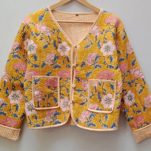 Könnte beinhalten: Gelbe Steppjacke mit einem Blumenmuster in Rosa und Blau. Die Jacke hat zwei Vordertaschen und einen V-Ausschnitt. Das Futter und die Paspeln sind aus hellorange-weiß gestreiftem Stoff. Die Jacke hängt an einem Holzkleiderbügel.
