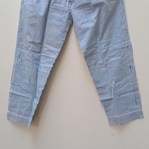 Peut inclure: Pantalon de pyjama à rayures bleues et blanches avec une taille à cordon de serrage. Le pantalon a une coupe décontractée et est fait d'un tissu léger. Les poignets sont bordés d'une fine bordure rose. Le pantalon est suspendu à un cintre en bois.