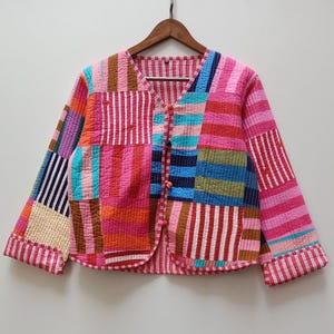 Puede incluir: Chaqueta patchwork colorida con cuello en V y mangas largas. La chaqueta presenta varios parches de tela en tonos rosa, rojo, azul, naranja y blanco. Tiene cierre de botones y está colgada en una percha de madera.