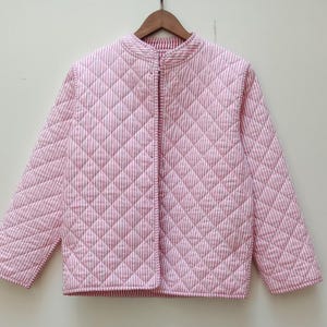 May include: A pink and white striped quilted jacket with a stand-up collar. The jacket is hanging on a wooden hanger. The jacket has a diamond pattern and a full-length zipper. The jacket is approximately 25 inches long.