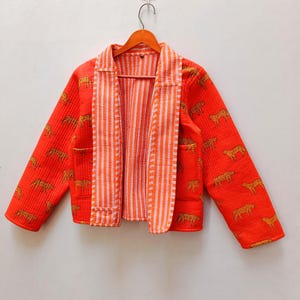 Puede incluir: Chaqueta naranja de frente abierto con textura acolchada y un patrón repetido de guepardos dorados. La chaqueta tiene un forro a rayas naranja y blanco y dos bolsillos delanteros. La chaqueta está colgada de una percha de madera.