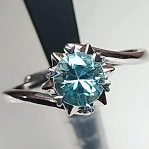 Peut inclure: Une bague en argent ornée d'une pierre précieuse ronde bleu clair. La pierre est entourée d'un motif en forme d'éclat d'étoile. Le corps de la bague épouse la pierre, créant un look moderne et élégant. La bague est posée sur fond blanc.