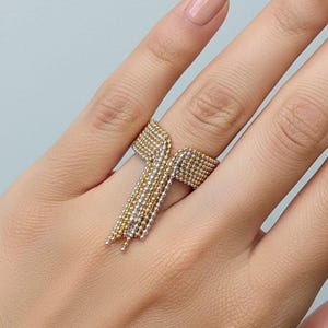 Pode incluir: Um anel de contas de ouro e prata com um design único. O anel apresenta uma série de pequenas contas dispostas em um padrão geométrico, com uma franja de contas em cascata. O anel é usado em uma mão.