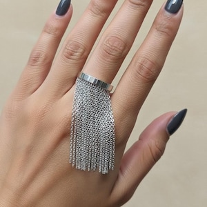 May include: A silver ring with a rectangular band and a cascade of dangling chains. The ring is worn on a finger, and the hand has dark gray nail polish. The jewelry is a statement piece.