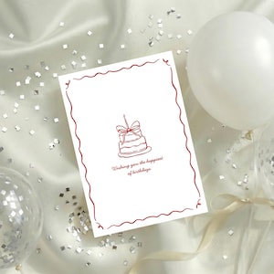 May include: A white birthday card with a red border and a cake illustration. The card reads "Wishing you the happiest of birthdays." The card is on a white satin surface with confetti and balloons.