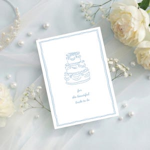 May include: A white card with a blue outline and a drawing of a tiered cake. The text "for the beautiful bride to be" is below the cake. The card is surrounded by pearls, flowers, and a tiara.