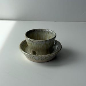 May include: A ceramic teacup and saucer set. The cup and saucer are a speckled beige and brown colour. The cup has a rounded shape and sits on a matching saucer with a slightly raised edge. The set is designed for serving tea or coffee.