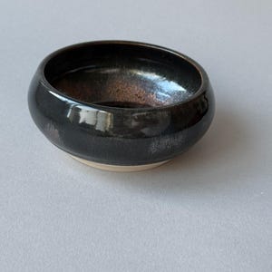 May include: A small, round ceramic bowl with a glossy black glaze on the exterior and interior. The interior features a subtle, textured copper-toned metallic finish at the base. The bottom edge of the bowl reveals the unglazed, light beige clay.