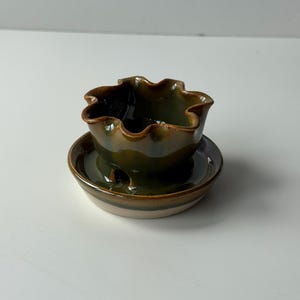 May include: A handmade ceramic bowl and saucer set. The bowl has a ruffled edge and a dark green glaze with brown accents. The saucer is a matching dark green with a cream-coloured base. The set is designed for serving small portions.